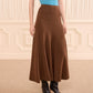 A person wearing a sleeveless blue top, black boots, and the Ulla Johnson Azaria Knit Skirt—a high-waisted brown skirt in soft merino wool—stands indoors against a cream-colored, soft paneled wall.