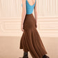 A person wearing a sleeveless blue top, long brown Azaria Knit Skirt by Ulla Johnson crafted from merino wool, and dark brown heeled boots, walking indoors on beige carpet with cream-colored soft panels lining the walls.