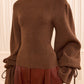 A person wearing the Ulla Johnson Camille Knit Pullover—a high-neck, ribbed turtleneck sweater featuring elongated balloon sleeves and drawstring cuffs—paired with high-waisted brown pants.