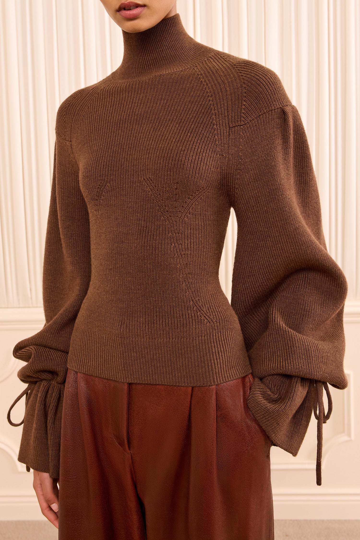 A person wearing the Ulla Johnson Camille Knit Pullover—a high-neck, ribbed turtleneck sweater featuring elongated balloon sleeves and drawstring cuffs—paired with high-waisted brown pants.