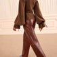 Wearing the Ulla Johnson Camille Knit Pullover—featuring a high neck and voluminous, elongated sleeves—a person pairs it with shiny brown leather pants while walking indoors on a beige carpet.