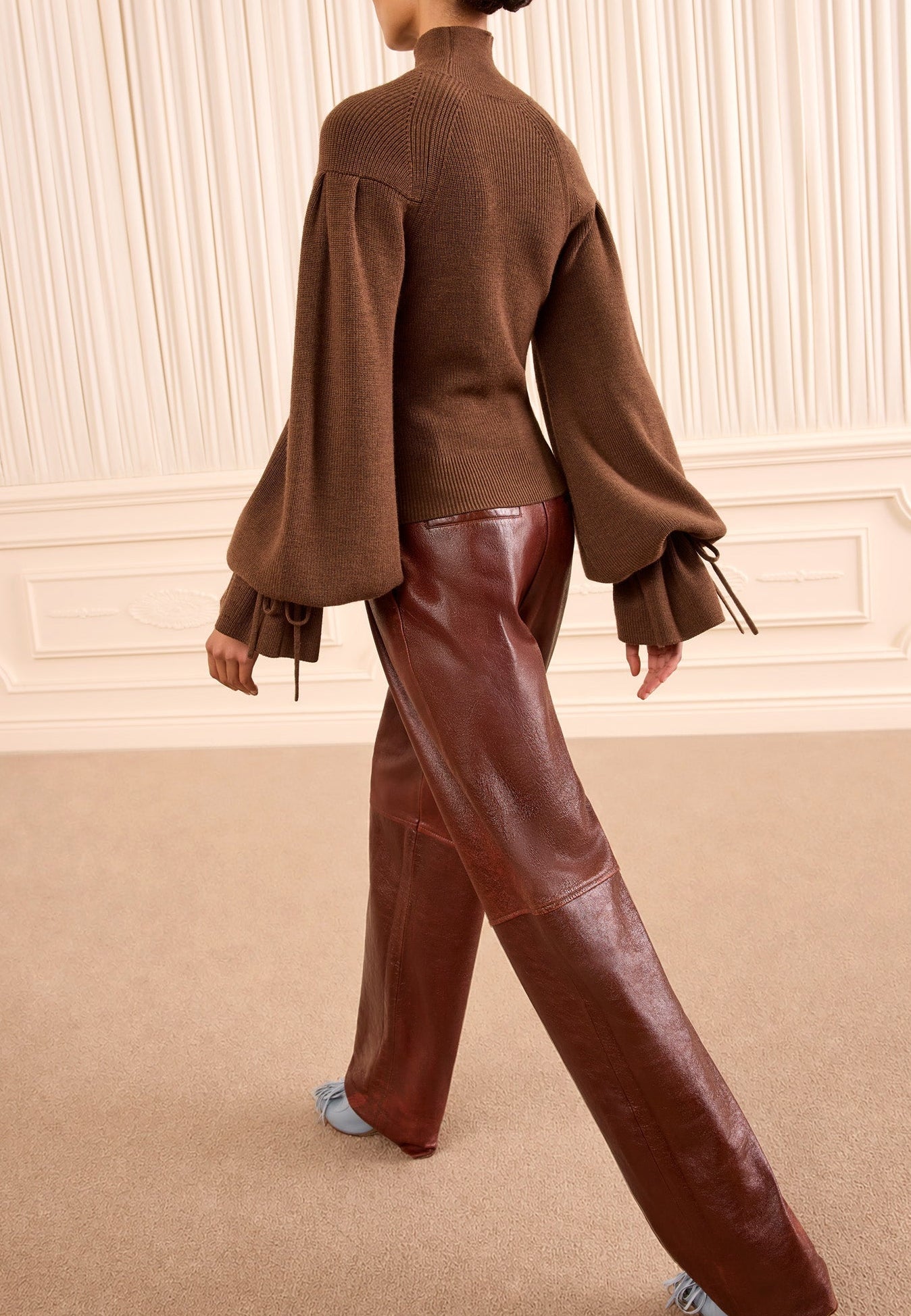 Wearing the Ulla Johnson Camille Knit Pullover—featuring a high neck and voluminous, elongated sleeves—a person pairs it with shiny brown leather pants while walking indoors on a beige carpet.
