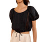 A woman wears the Ulla Johnson Clara Top—a cropped black cotton blouse featuring short sleeves, button-front closure, and delicate lace details reminiscent of a Victorian lace top—paired with high-waisted cream shorts.