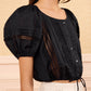 A woman wears the Ulla Johnson Clara Top—a cropped black cotton top with lace panels inspired by Victorian lace—paired with a white high-waisted skirt, standing indoors against a beige background.