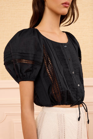 A woman wears the Ulla Johnson Clara Top—a cropped black cotton top with lace panels inspired by Victorian lace—paired with a white high-waisted skirt, standing indoors against a beige background.