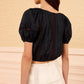 A woman wearing the Ulla Johnson Clara Top, a cropped black cotton style with lace panels, paired with a white high-waisted skirt and standing against a beige textured wall.