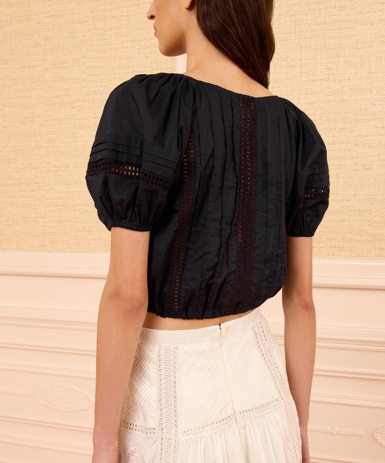 A woman wearing the Ulla Johnson Clara Top, a cropped black cotton style with lace panels, paired with a white high-waisted skirt and standing against a beige textured wall.