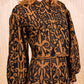 A person stands before light pleated curtains, wearing a long-sleeved dress with a black and brown abstract pattern, paired with the Ulla Johnson Coletta Applique Jacket.