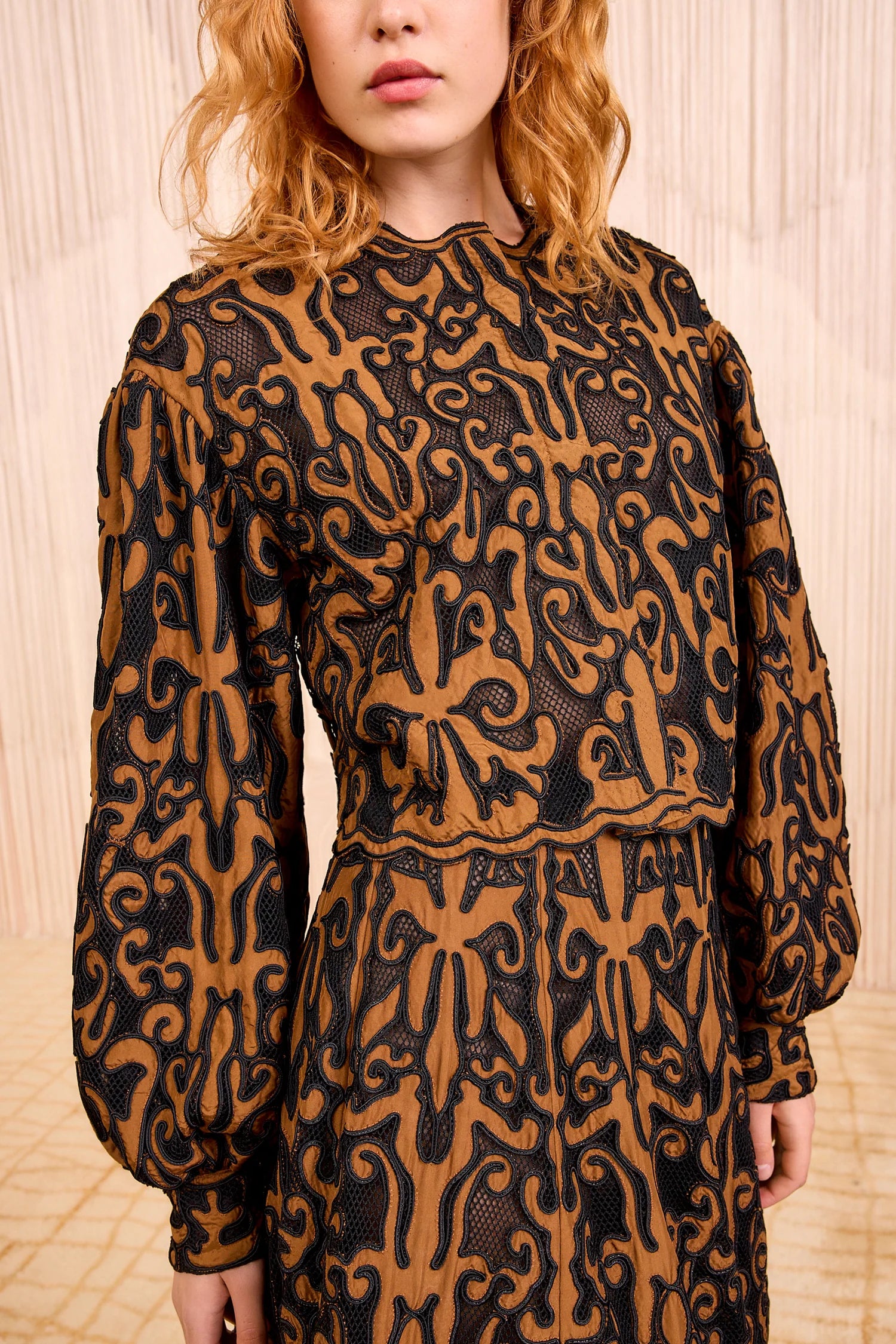 A person stands before light pleated curtains, wearing a long-sleeved dress with a black and brown abstract pattern, paired with the Ulla Johnson Coletta Applique Jacket.