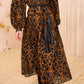 A woman stands in a neutral-toned room wearing a long-sleeve, calf-length brown dress with black patterns, accented by the Ulla Johnson Coletta Applique Jacket draped over her shoulders, a matching tassel belt, and black boots.