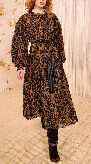 A woman stands in a neutral-toned room wearing a long-sleeve, calf-length brown dress with black patterns, accented by the Ulla Johnson Coletta Applique Jacket draped over her shoulders, a matching tassel belt, and black boots.