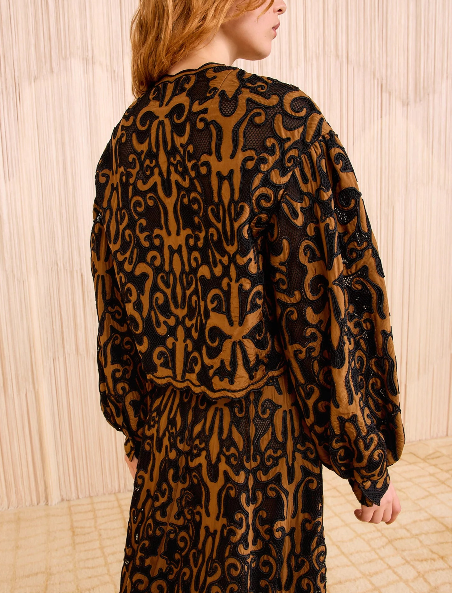 A person with light brown hair wears the Ulla Johnson Coletta Applique Jacket, a black and gold patchwork piece with long sleeves, standing before a beige textured background.