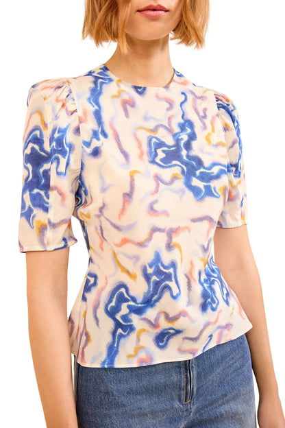 Woman wearing the Ulla Johnson Corine Silk Top, a short-sleeve blouse with a colorful abstract swirl pattern, paired with light blue wide-leg pants.