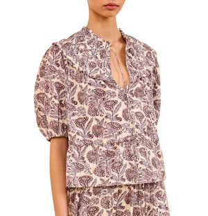 Woman wearing the Ulla Johnson Elvie Top, a lightweight cotton-blend short-sleeve blouse with a ruffled collar, keyhole neckline, and purple and beige floral batik print.