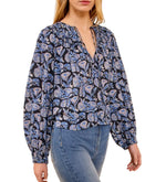 A woman wears the Ulla Johnson Florelle Blouse, a long-sleeve cotton top with shadow stripes and ikat-inspired blue and black florals, featuring a voluminous fit, styled with blue jeans.