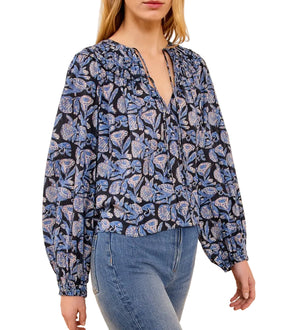 A woman wears the Ulla Johnson Florelle Blouse, a long-sleeve cotton top with shadow stripes and ikat-inspired blue and black florals, featuring a voluminous fit, styled with blue jeans.