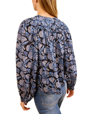 A person in the Ulla Johnson Florelle Blouse, featuring a voluminous fit with blue and black ikat-inspired florals, paired with blue jeans and viewed from the back.
