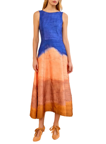 A woman wears the Ulla Johnson Georgia Satin Midi Dress, a sleeveless, open-back style in crinkled satin with a blue bodice fading into peach and copper, paired with tan ribbon-tied shoes.