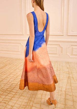 A woman stands in a beige-carpeted room wearing the Ulla Johnson Georgia Satin Midi Dress, featuring a blue, peach, and orange gradient pattern, paired with tan sandals.