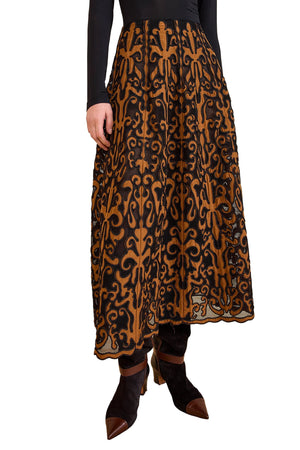 A person wearing the Gili Midi Skirt by Ulla Johnson, featuring black mesh with intricate brown embroidery, paired with a black long-sleeve top and black pointed shoes.