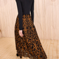 A woman stands on a beige patterned floor, facing away from the camera, wearing a black turtleneck top and the Ulla Johnson Gili Midi Skirt featuring black embroidered patterns and Victorian-inspired design.