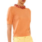 A person shown from shoulders to hips wears the Ulla Johnson Jude Embellished Top—an embellished, peach merino wool short-sleeve sweater—paired with yellow pants and a chunky coral floral necklace.