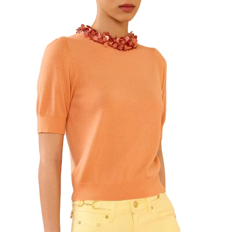 A person shown from shoulders to hips wears the Ulla Johnson Jude Embellished Top—an embellished, peach merino wool short-sleeve sweater—paired with yellow pants and a chunky coral floral necklace.