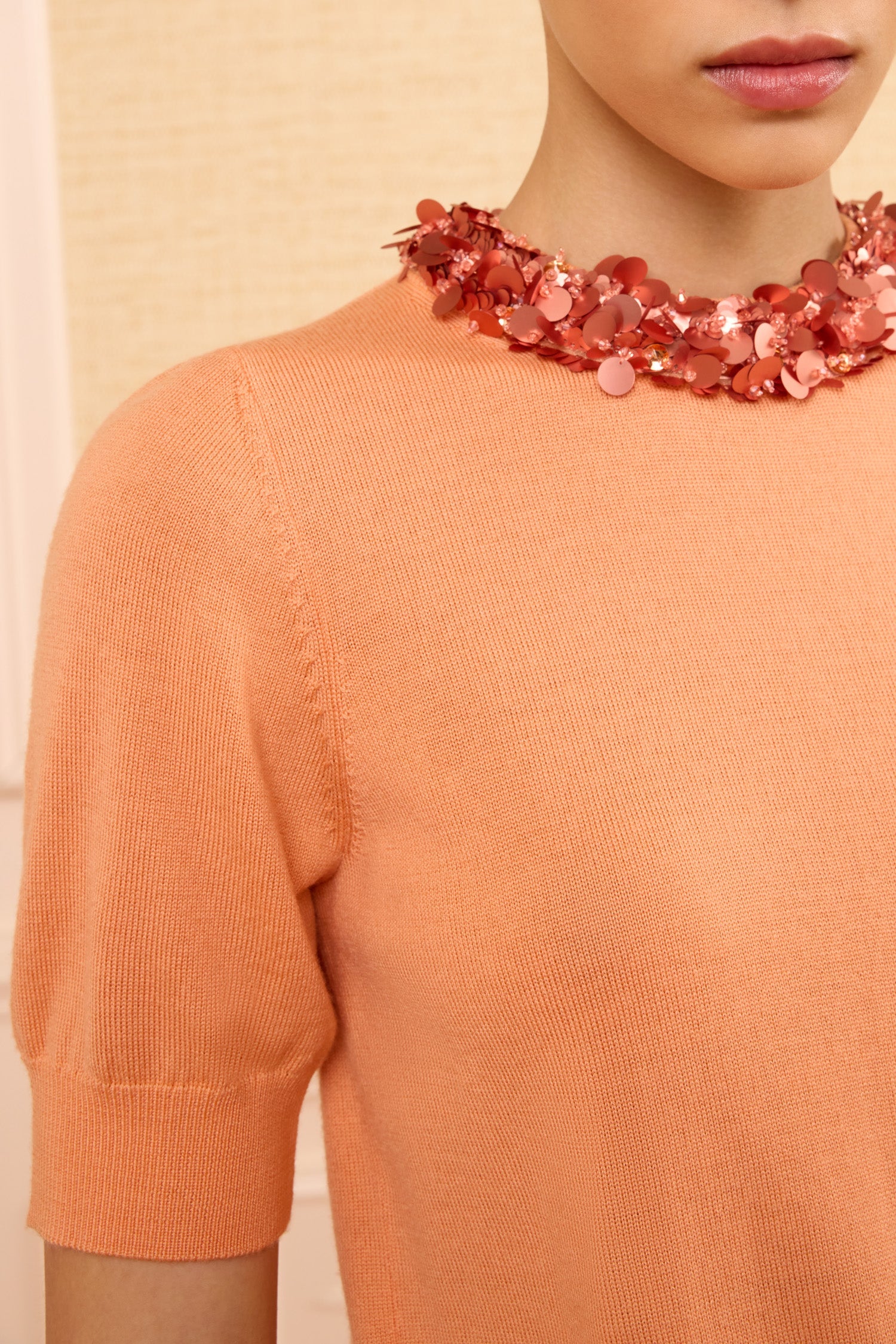 Model wears the Ulla Johnson Jude Embellished Top in peach with short sleeves, paired with a chunky pink sequin necklace, shown from shoulders to mouth against a neutral background.