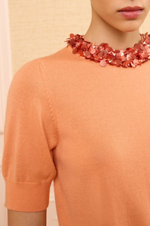 Model wears the Ulla Johnson Jude Embellished Top in peach with short sleeves, paired with a chunky pink sequin necklace, shown from shoulders to mouth against a neutral background.