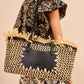 A woman in a black and beige floral outfit carries the Ulla Johnson Lali Small East West Tote, a hand-woven bag featuring black leather accents and decorative straw detailing.