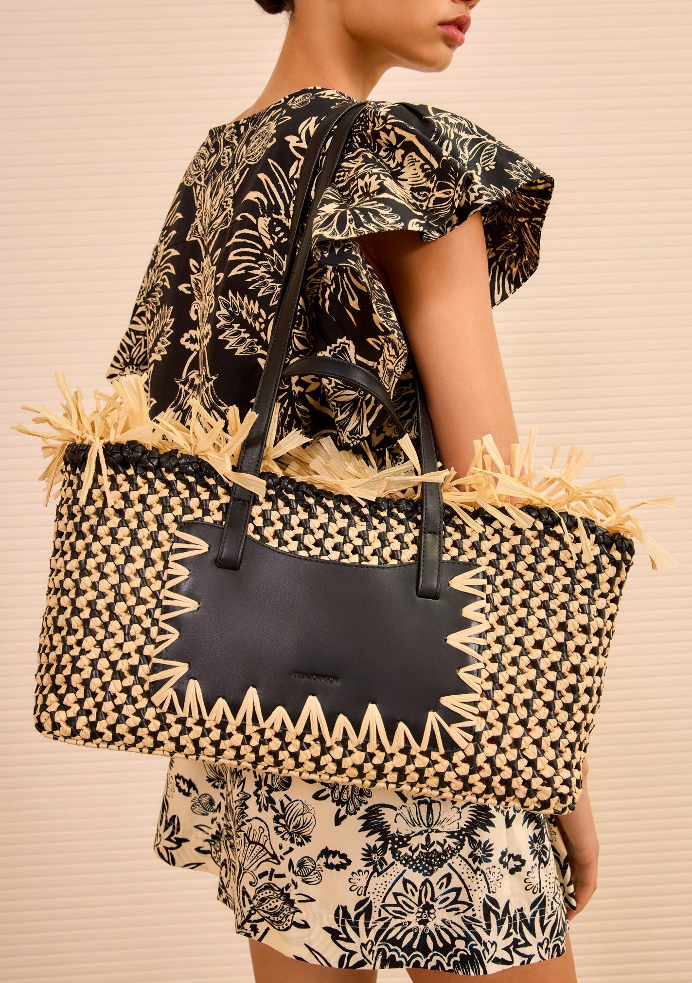 A woman in a black and beige floral outfit carries the Ulla Johnson Lali Small East West Tote, a hand-woven bag featuring black leather accents and decorative straw detailing.