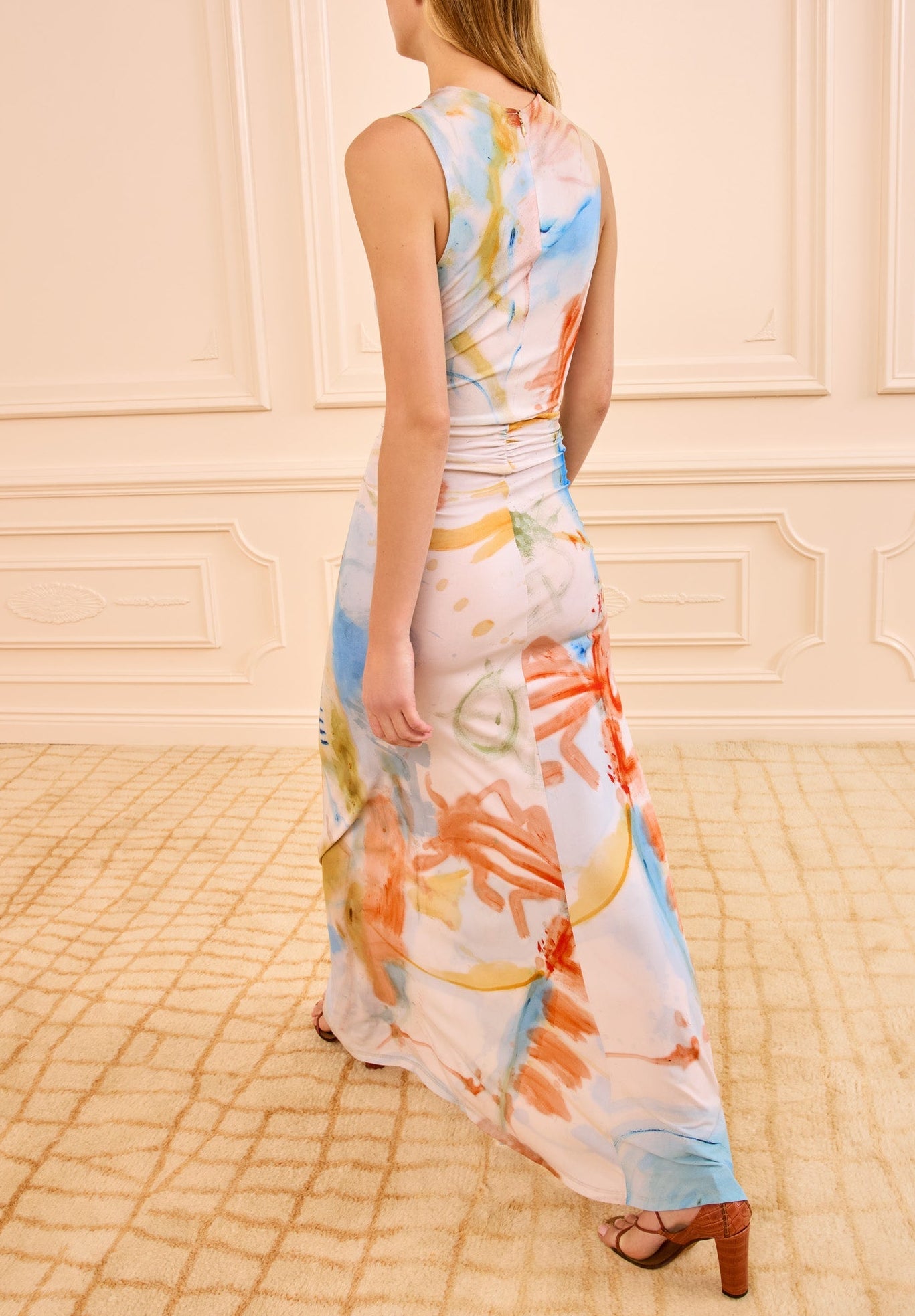 A woman in a sleeveless Ulla Johnson Lana Ruched Jersey Dress, inspired by colorful abstract art, walks on a beige carpet in a room with cream paneled walls.