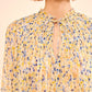 A person wearing the Ulla Johnson Leandra Silk Blouse, a light chiffon top with yellow, blue, and white floral pleats and a tied keyhole neckline.