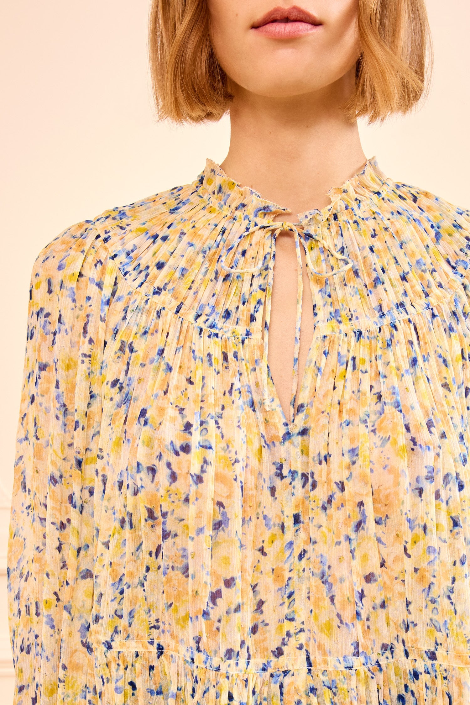 A person wearing the Ulla Johnson Leandra Silk Blouse, a light chiffon top with yellow, blue, and white floral pleats and a tied keyhole neckline.