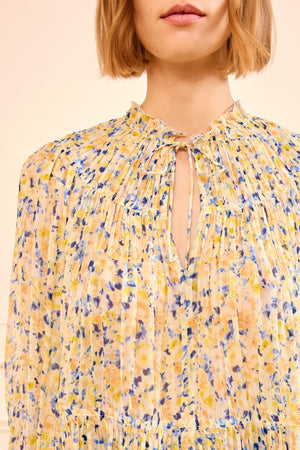 A person wearing the Ulla Johnson Leandra Silk Blouse, a light chiffon top with yellow, blue, and white floral pleats and a tied keyhole neckline.