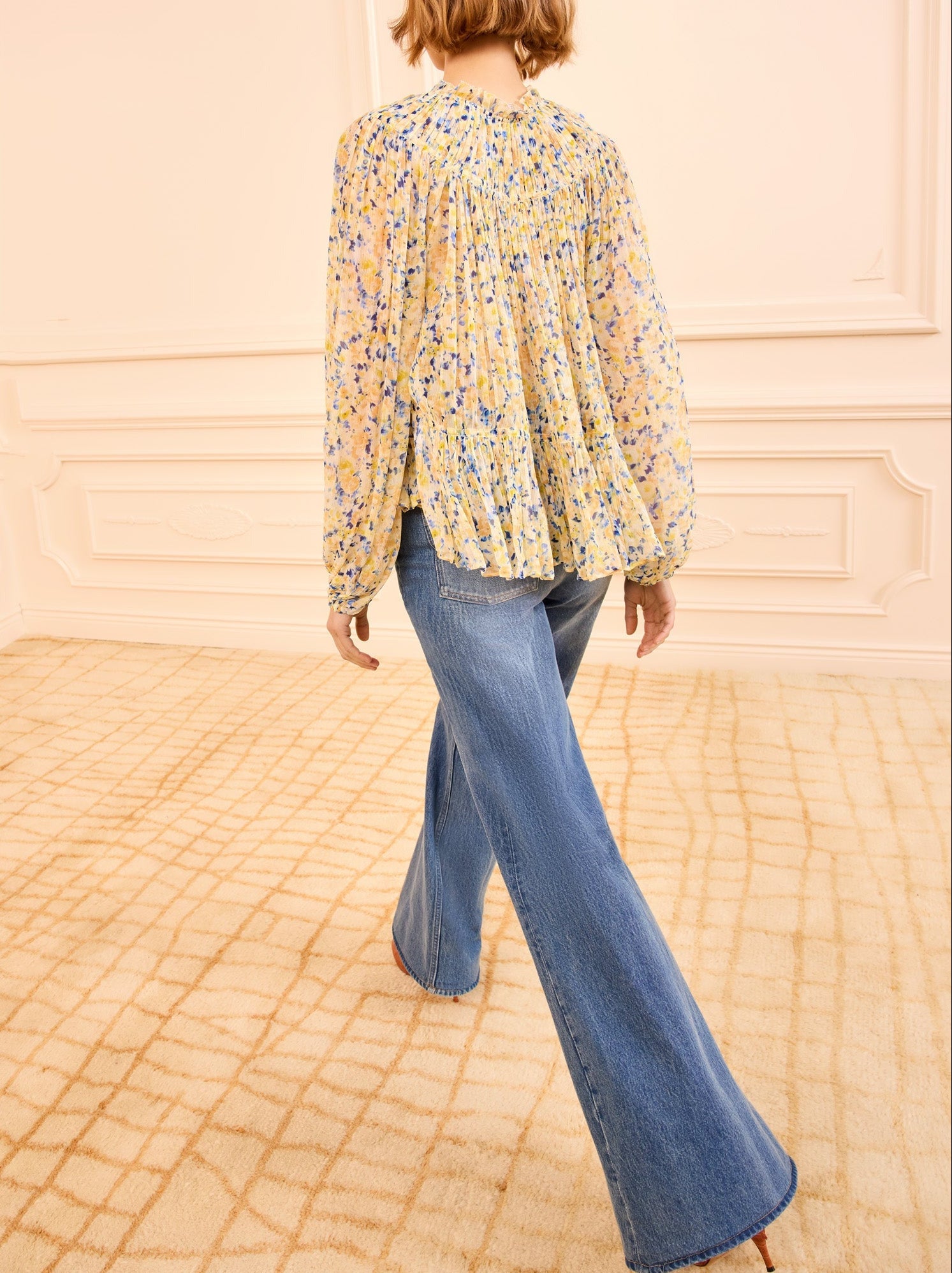 A person wearing a light floral Leandra Silk Blouse by Ulla Johnson and flared blue jeans walks away in a beige carpeted room with cream paneled walls.