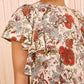 A woman wears the Ulla Johnson Lena Flutter Sleeve Blouse, cream-colored silk crepe de chine with red, green, and orange floral patterns and ruffled short sleeves.