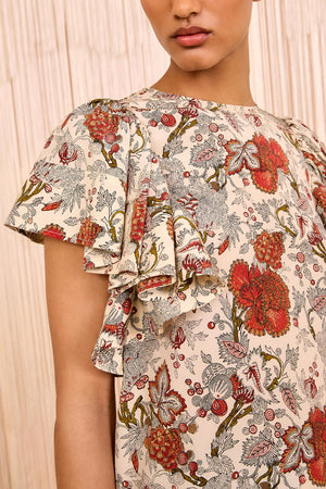 A woman wears the Ulla Johnson Lena Flutter Sleeve Blouse, cream-colored silk crepe de chine with red, green, and orange floral patterns and ruffled short sleeves.