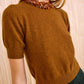 A woman wears the Ulla Johnson Liv Embroidered Cashmere Knit in umber brown with a chunky beaded collar, paired with dark green pants, standing against a beige textured background.