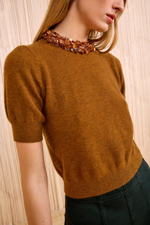 A woman wears the Ulla Johnson Liv Embroidered Cashmere Knit in umber brown with a chunky beaded collar, paired with dark green pants, standing against a beige textured background.