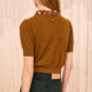 A woman stands with her back to the camera wearing the Ulla Johnson Liv Embroidered Cashmere Knit, an umber brown short-sleeve top, paired with dark green pants and a beaded collar visible around her neck.