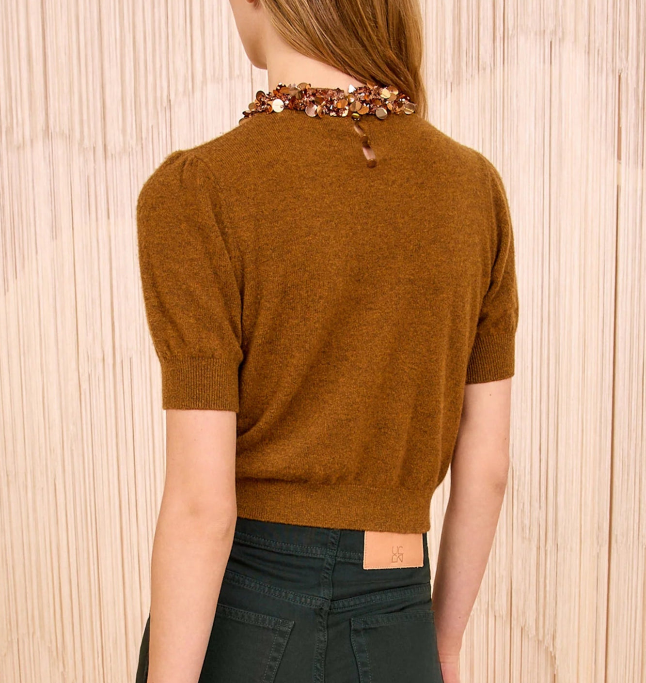 A woman stands with her back to the camera wearing the Ulla Johnson Liv Embroidered Cashmere Knit, an umber brown short-sleeve top, paired with dark green pants and a beaded collar visible around her neck.