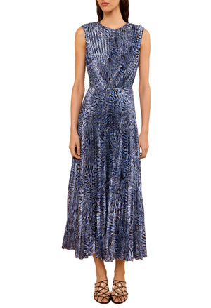 A woman in the Ulla Johnson Malin Satin Dress—a sleeveless, ankle-length blue and black patterned pleated midi—stands against a white background wearing black strappy shoes.