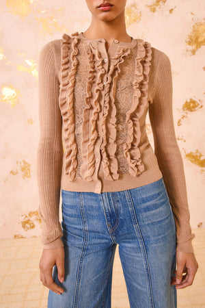 A person wearing the Ulla Johnson Maryel Cardigan, a beige, long-sleeve merino wool piece with ruffle and lace details, paired with blue high-waisted jeans.