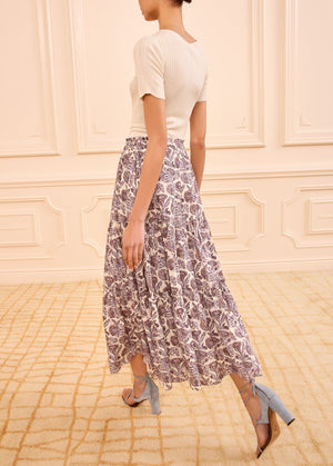 A woman walks indoors on a carpeted floor, wearing a short-sleeve white ribbed top, light blue block-heeled sandals, and the Nessa Skirt by Ulla Johnson—a flowy cotton-blend skirt with batik-inspired florals and an elastic waist.