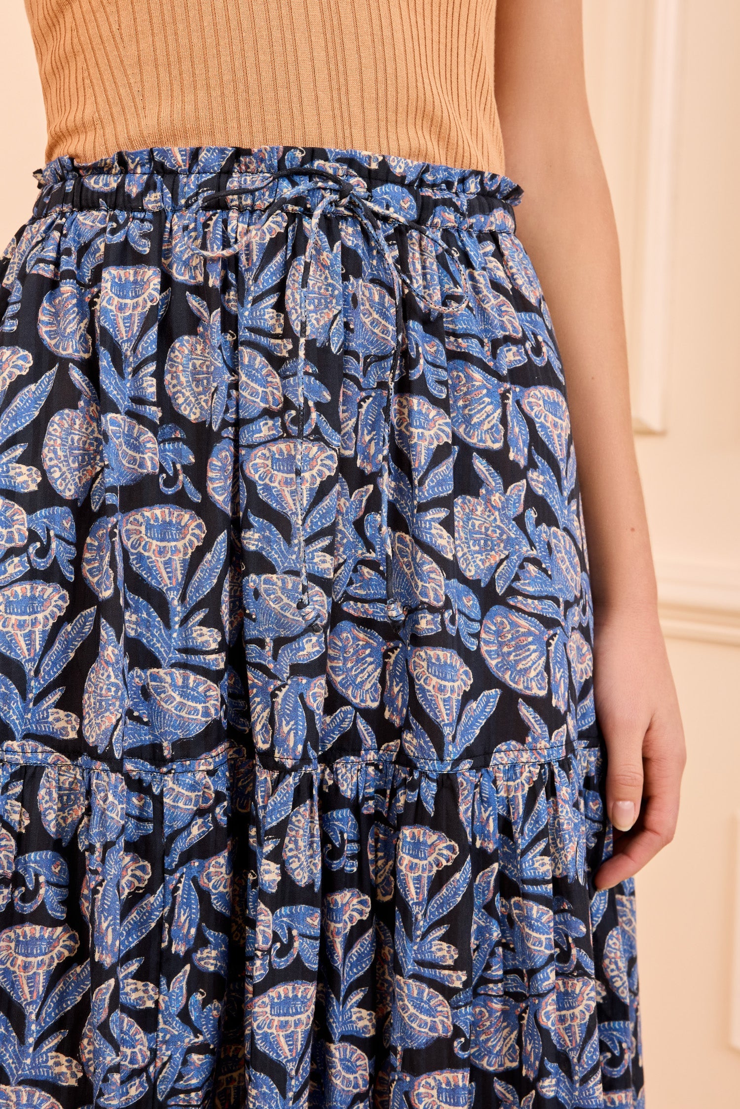A person wears a sleeveless, ribbed tan top paired with the Ulla Johnson Nessa Skirt—a high-waisted, cotton-blend piece featuring batik-inspired florals.