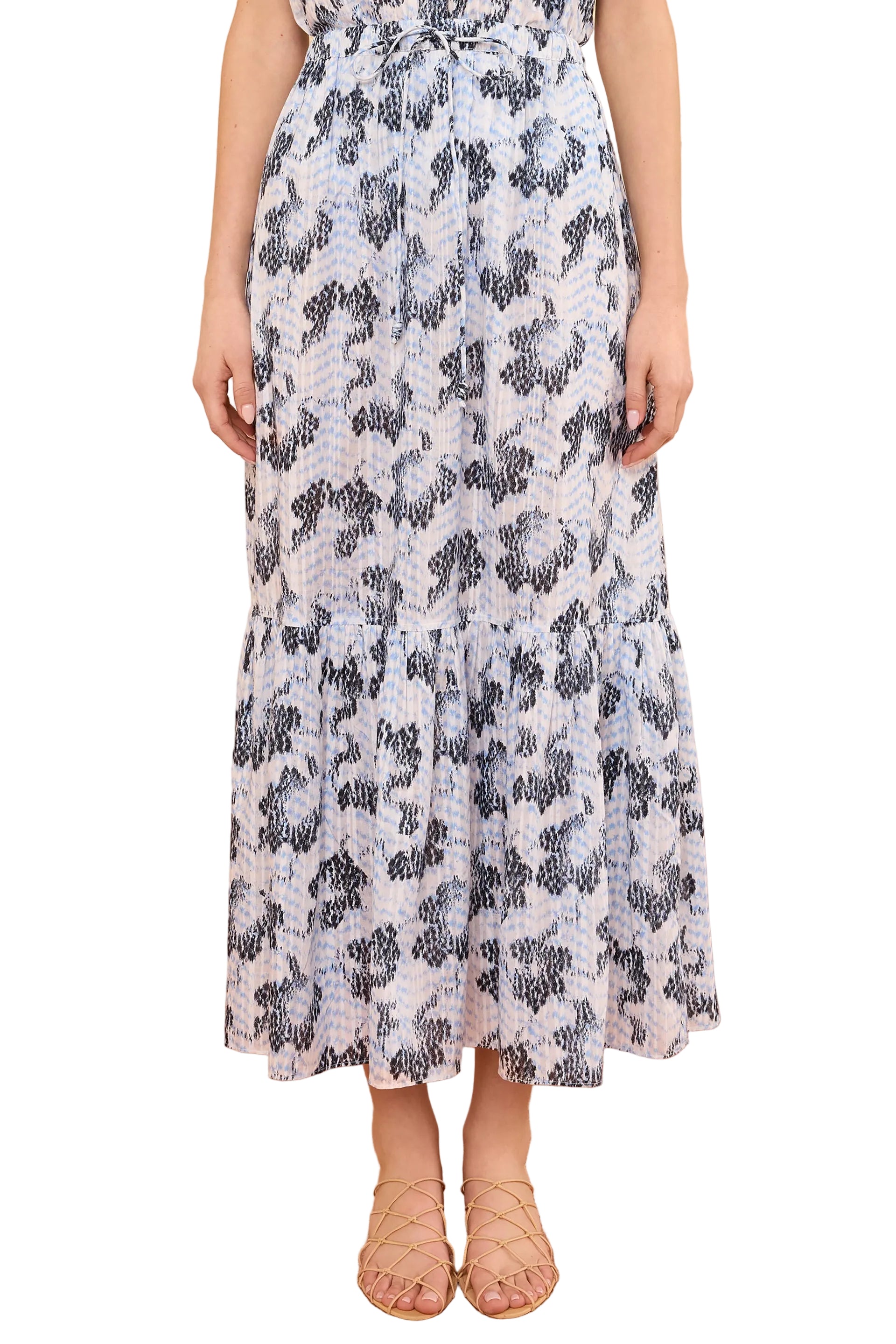 A person wearing the Ulla Johnson Olina Midi Skirt, a light-colored cotton-blend skirt with blue and black abstract print, styled with tan strappy sandals.