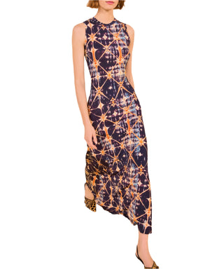 A woman wears the Ulla Johnson Orla Dress, a sleeveless maxi featuring geometric shibori textures in dark blue, orange, and white, paired with animal print shoes.