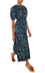 A person wearing the Ulla Johnson Othella Cotton Dress in dark blue with a mid-length floral design and short sleeves, paired with brown heeled loafers, walks forward.