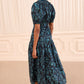 A woman with her hair in a bun wears the Othella Cotton Dress by Ulla Johnson—a dark blue floral print maxi dress with elegant puff sleeves—and black heeled shoes, standing in a beige room with decorative wall panels.